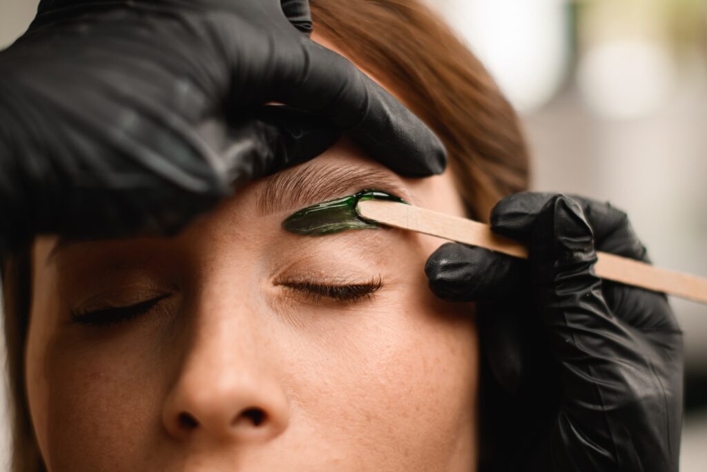 gloved hands applying green wax to an eyebrow using a wooden spatula while the person’s eyes are closed