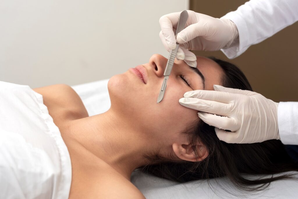 gloved practitioner holding a dermaplaning blade against a woman’s cheek while she lies on a treatment bed
