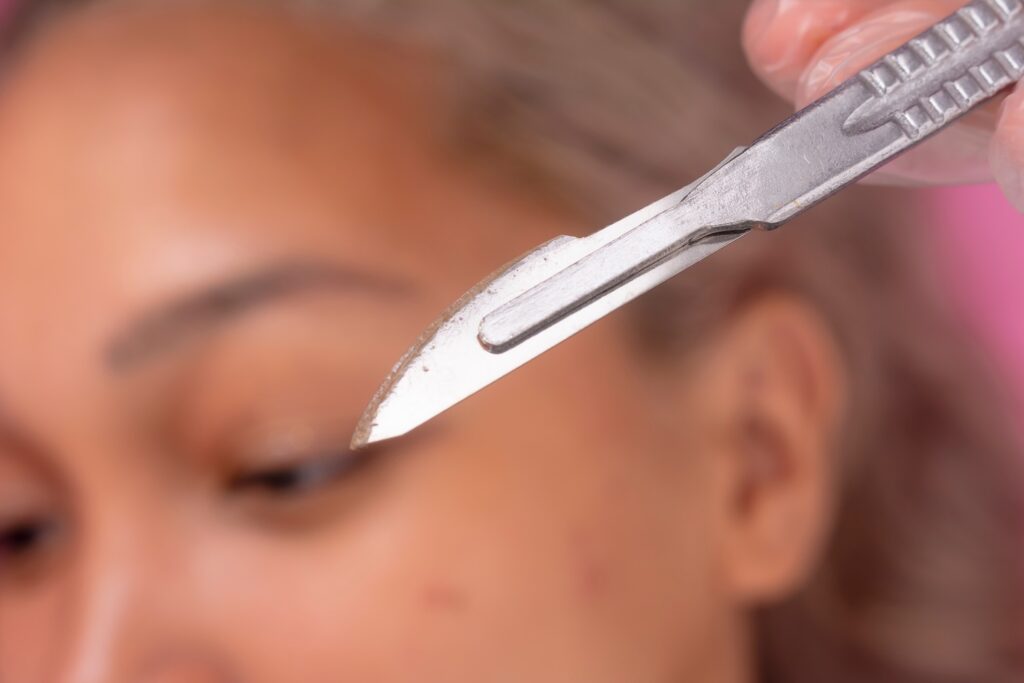 close-up of a dermaplaning blade being used near a woman’s eyebrow during a facial treatment