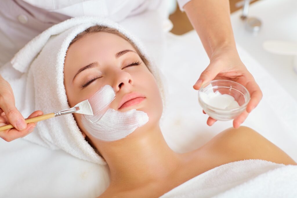 person lying on treatment bed while a brush applies a white facial mask to the cheek