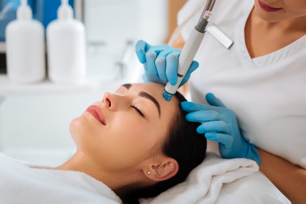 woman lying with eyes closed while a practitioner wearing blue gloves uses a handheld facial device on her forehead