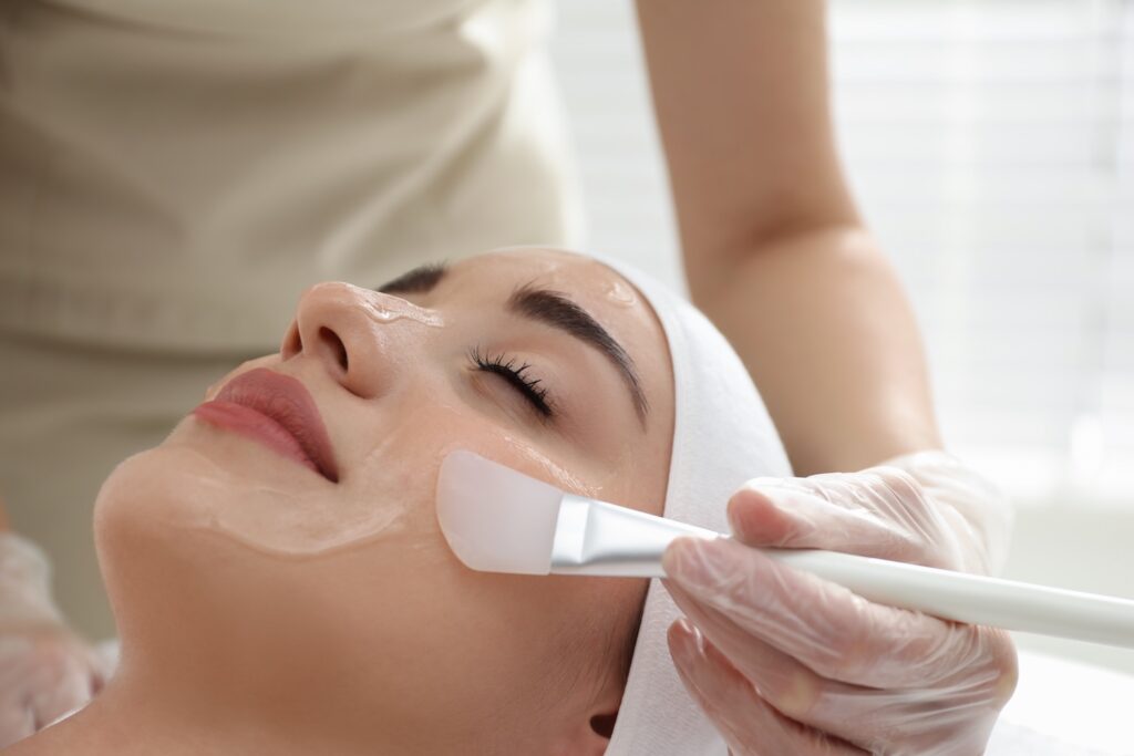 esthetician wearing gloves applies clear gel to a woman’s cheek using a flat facial applicator