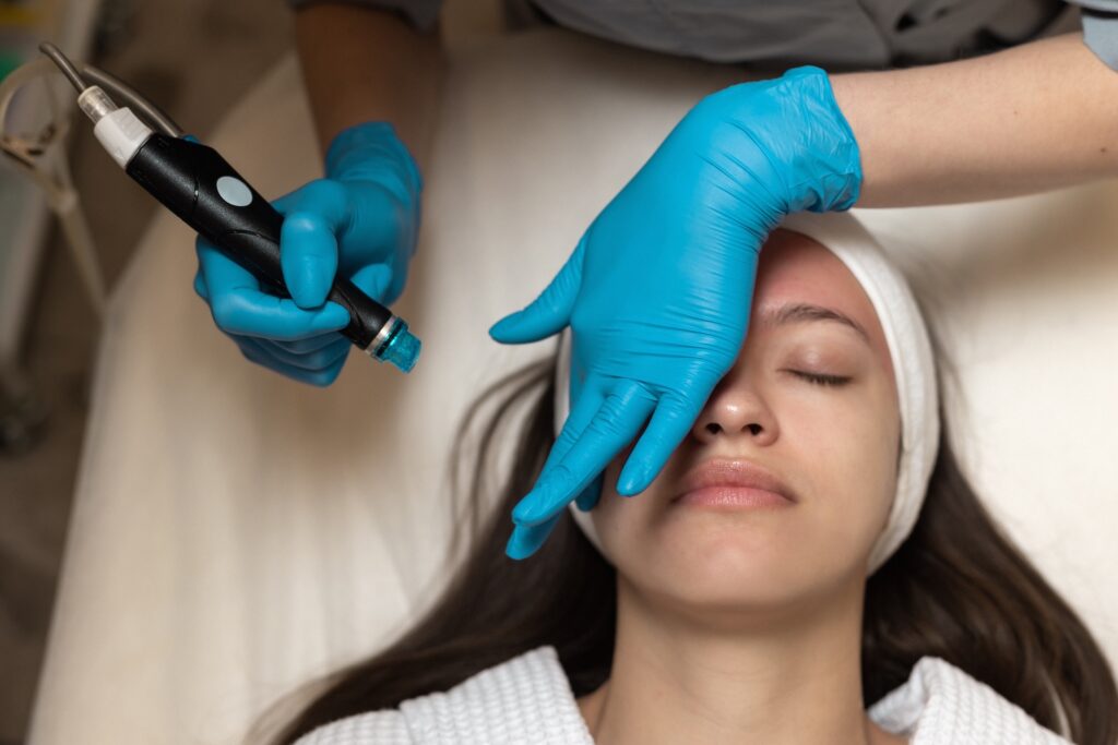 esthetician wearing blue gloves holds a handheld facial device near a woman’s face as she lies with eyes closed