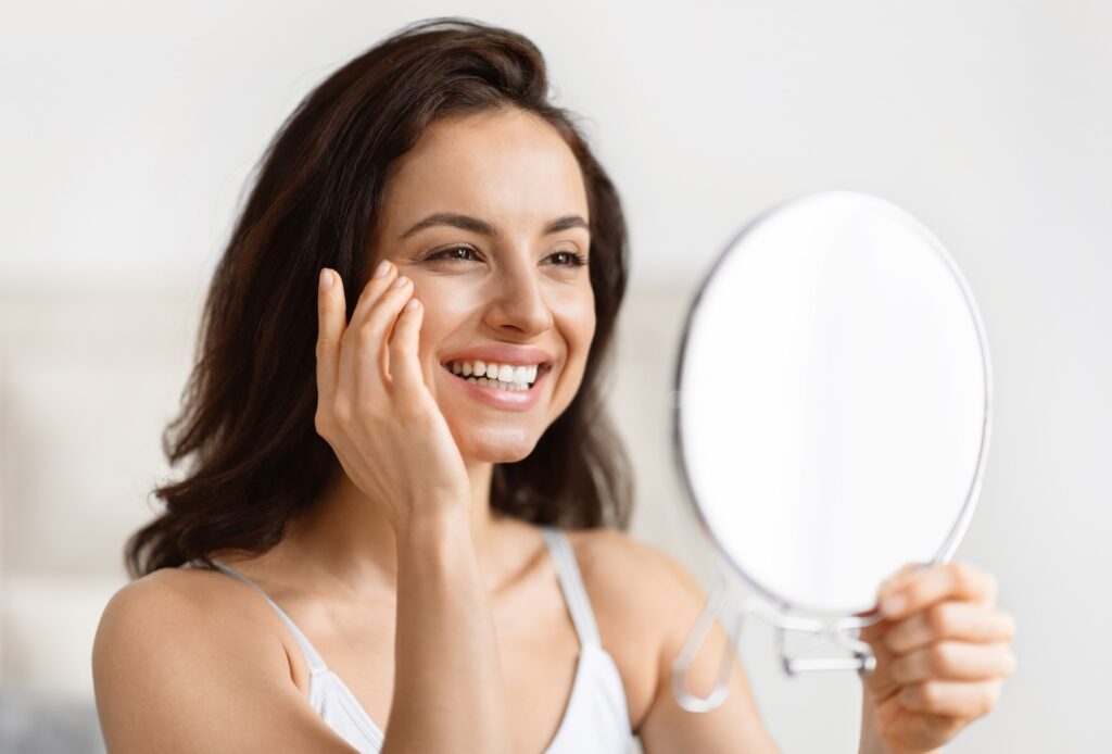 young woman smiling while examining her face in a handheld mirror indoors