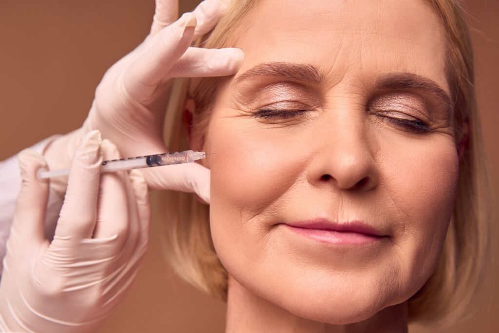 A gloved clinician holds a syringe near the cheek of a woman with her eyes closed.