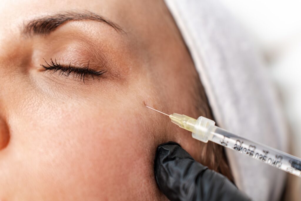 A gloved hand holds a syringe near the skin at the outer corner of a woman’s closed eye.