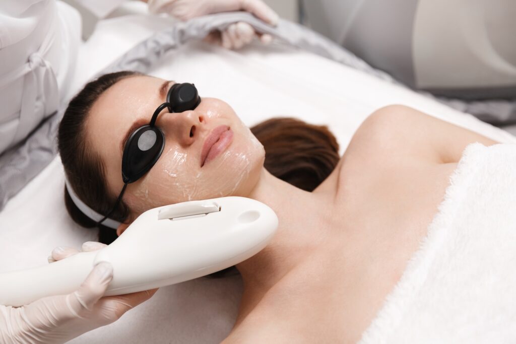 Woman lying down with eye shields and gel on her face while a technician holds a handheld laser device near her cheek.