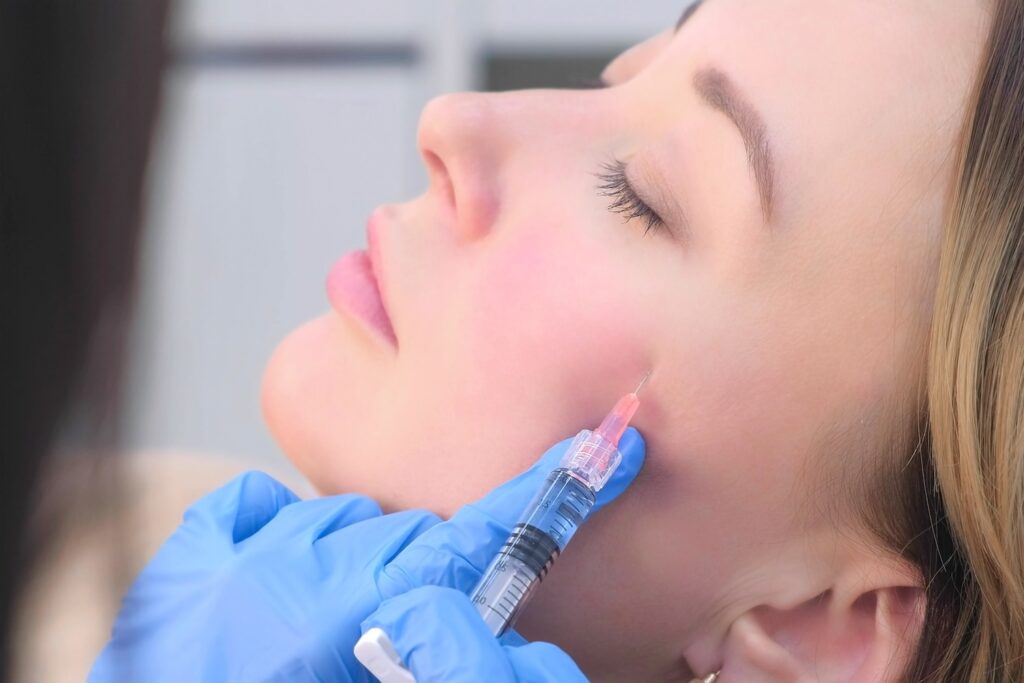 A clinician in blue gloves holds a syringe against a woman’s cheek as she lies with her eyes closed.