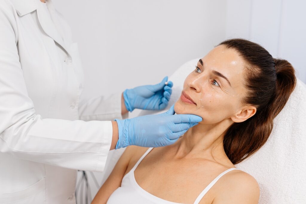 Provider wearing blue gloves examines a woman’s face with marking dots as she sits in a chair.