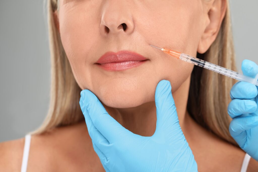 Gloved hands hold a syringe near a woman’s cheek while another hand supports her chin.