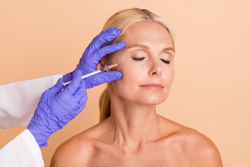 Practitioner wearing purple gloves holding a syringe near a woman’s cheek as she sits with her eyes closed.
