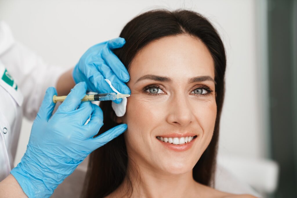A smiling woman receives an injection near the outer corner of her eye from a professional wearing blue medical gloves.
