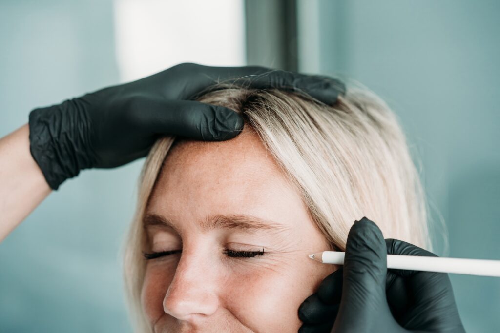 Practitioner in black gloves marking the outer corner of a woman’s closed eye with a white pencil during treatment preparation.
