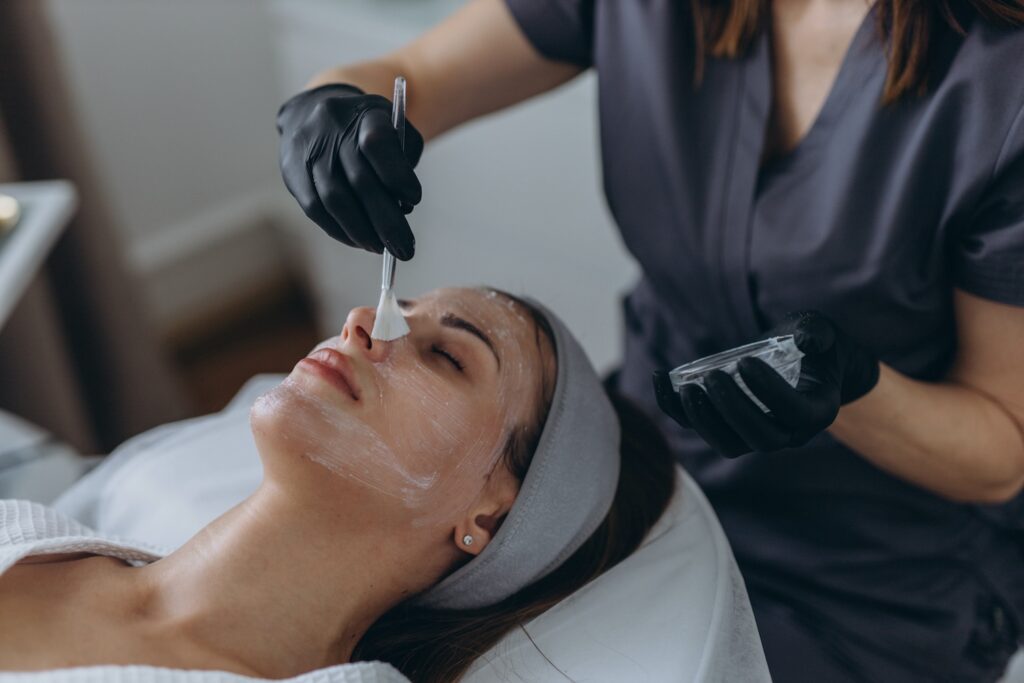 a person lying on a treatment bed with eyes closed while a provider applies a chemical peel to the face using a small brush