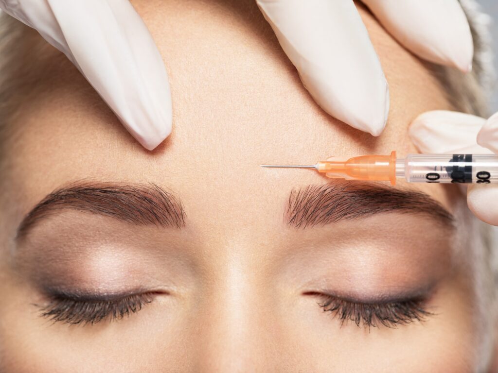gloved hands holding a syringe near a woman’s forehead above her eyebrow
