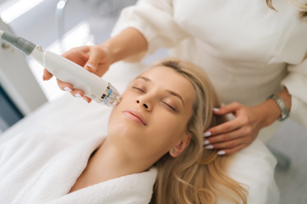 a person lying down with eyes closed receiving a facial microneedling treatment with a handheld device