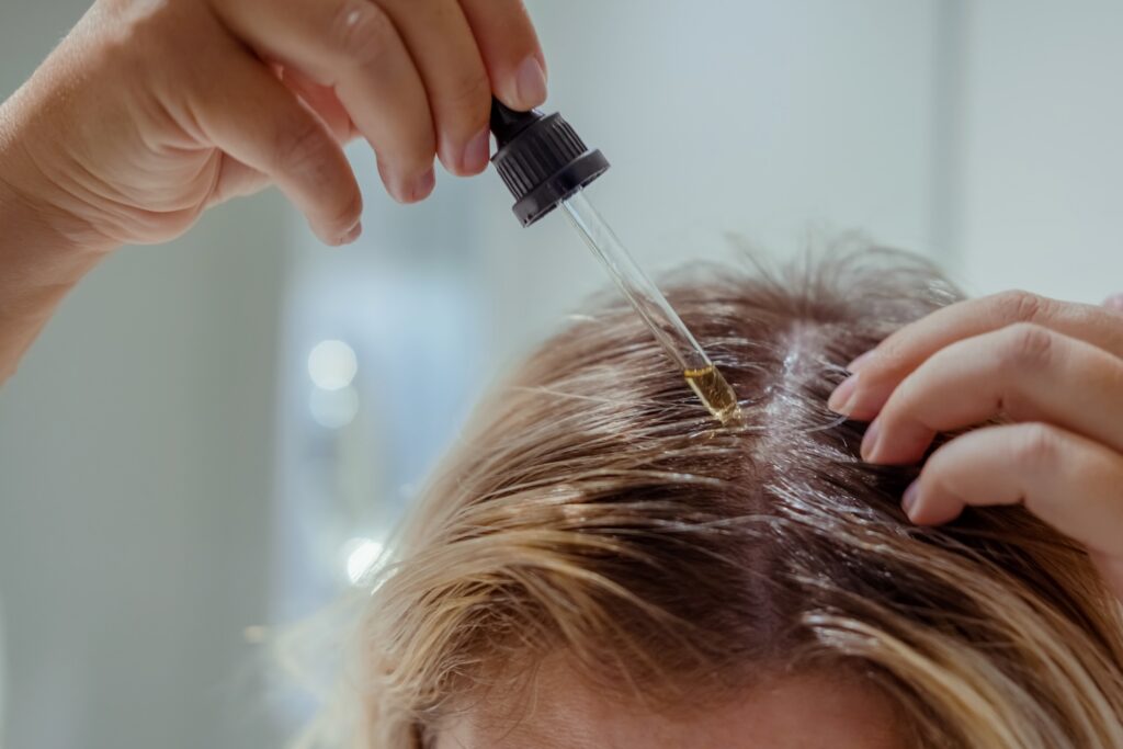 hand using a dropper to apply liquid treatment to scalp along a parted section of hair