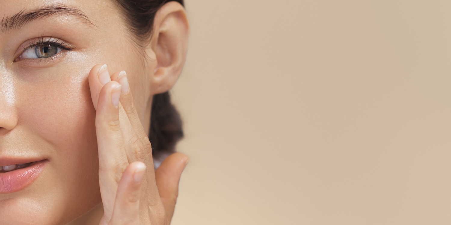 A woman gently touches her cheek with her fingers, shown in a partial face close-up against a neutral background.