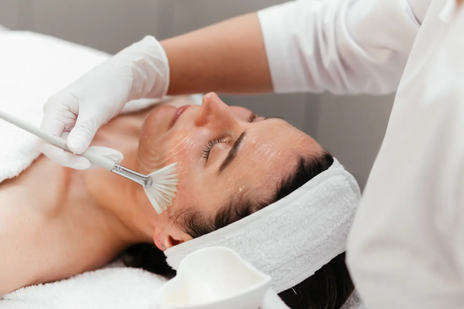 A practitioner wearing white gloves applies a clear product to a woman’s cheek with a fan brush while she lies on a treatment bed.