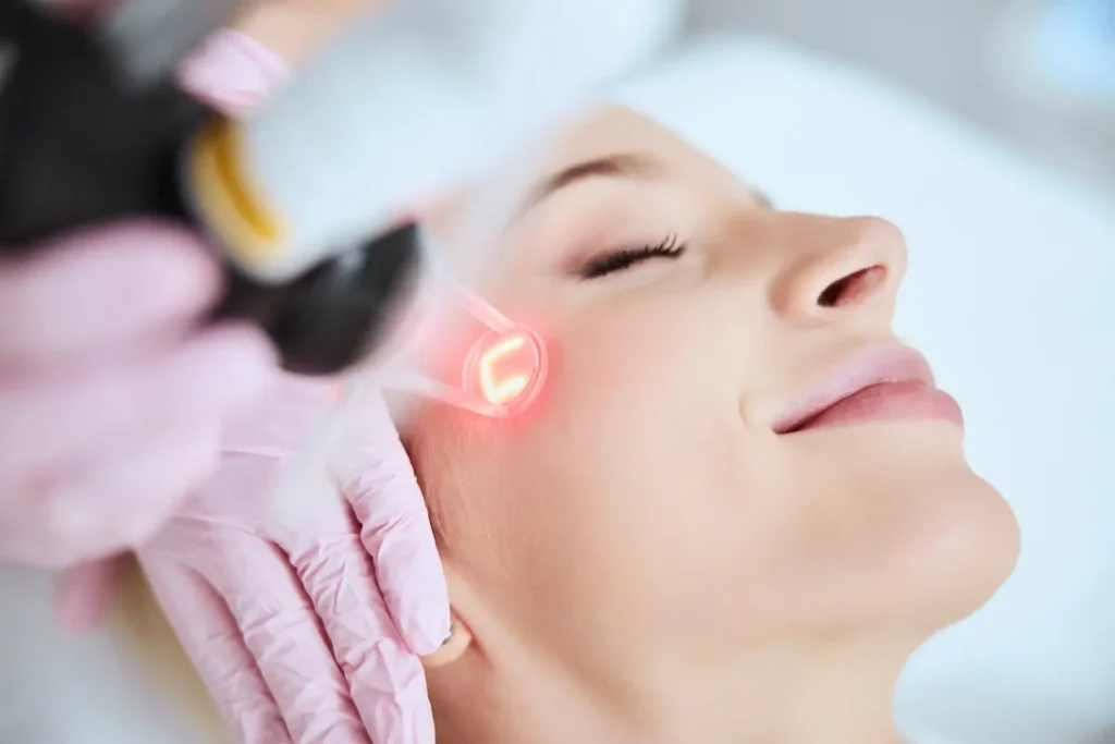 Gloved hand holds a handheld device emitting red light against a person’s cheek as they lie back with eyes closed