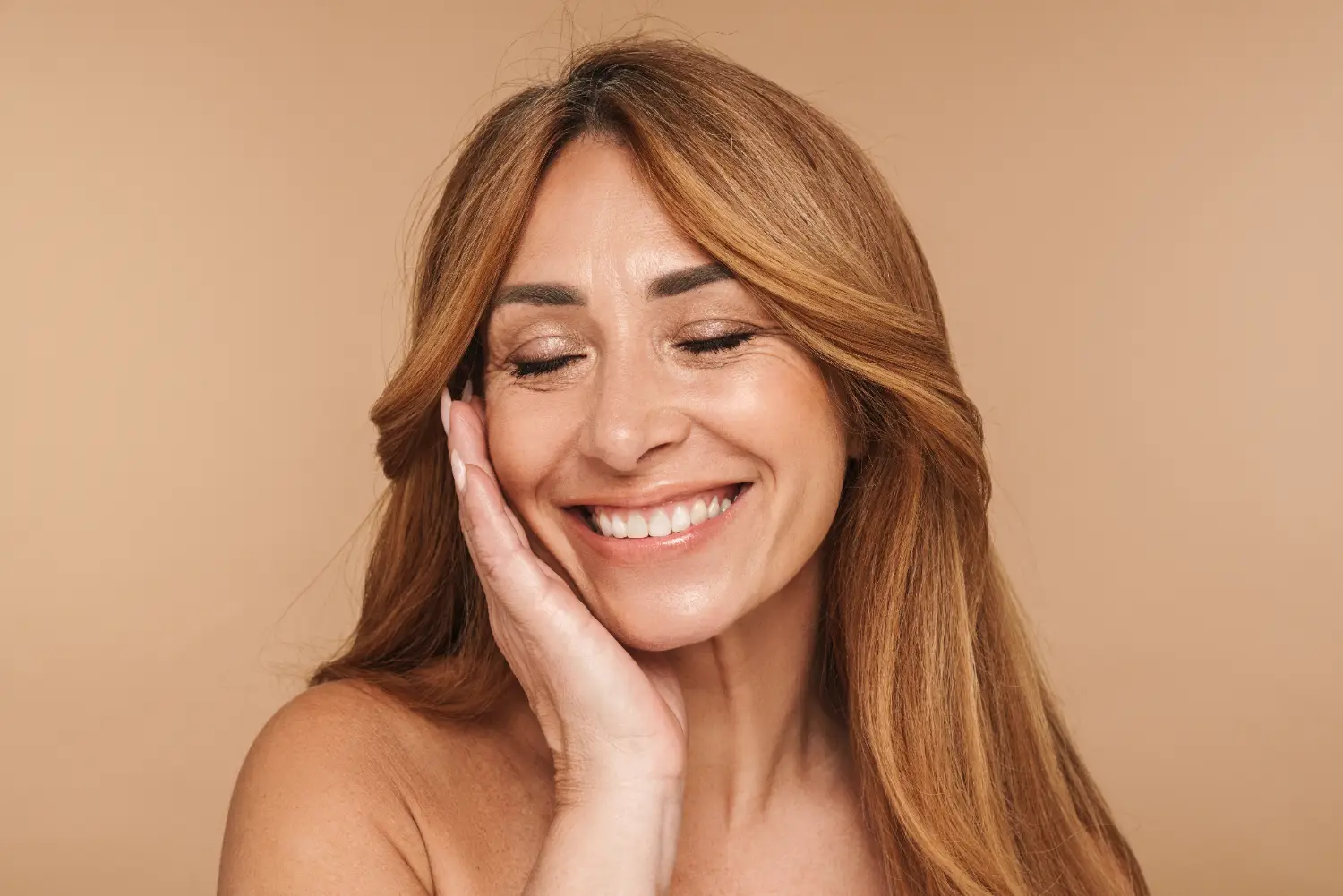 Close-up of a woman with long light brown hair smiling with her hand resting on her cheek against a beige background.