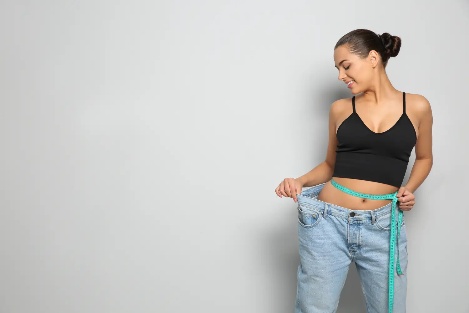 Smiling woman wearing oversized jeans while measuring her waist to show weight loss progress.