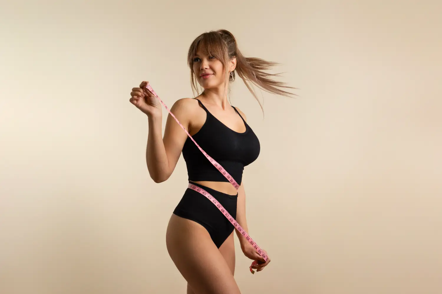 Fit woman in black activewear measuring her waist with a pink measuring tape.