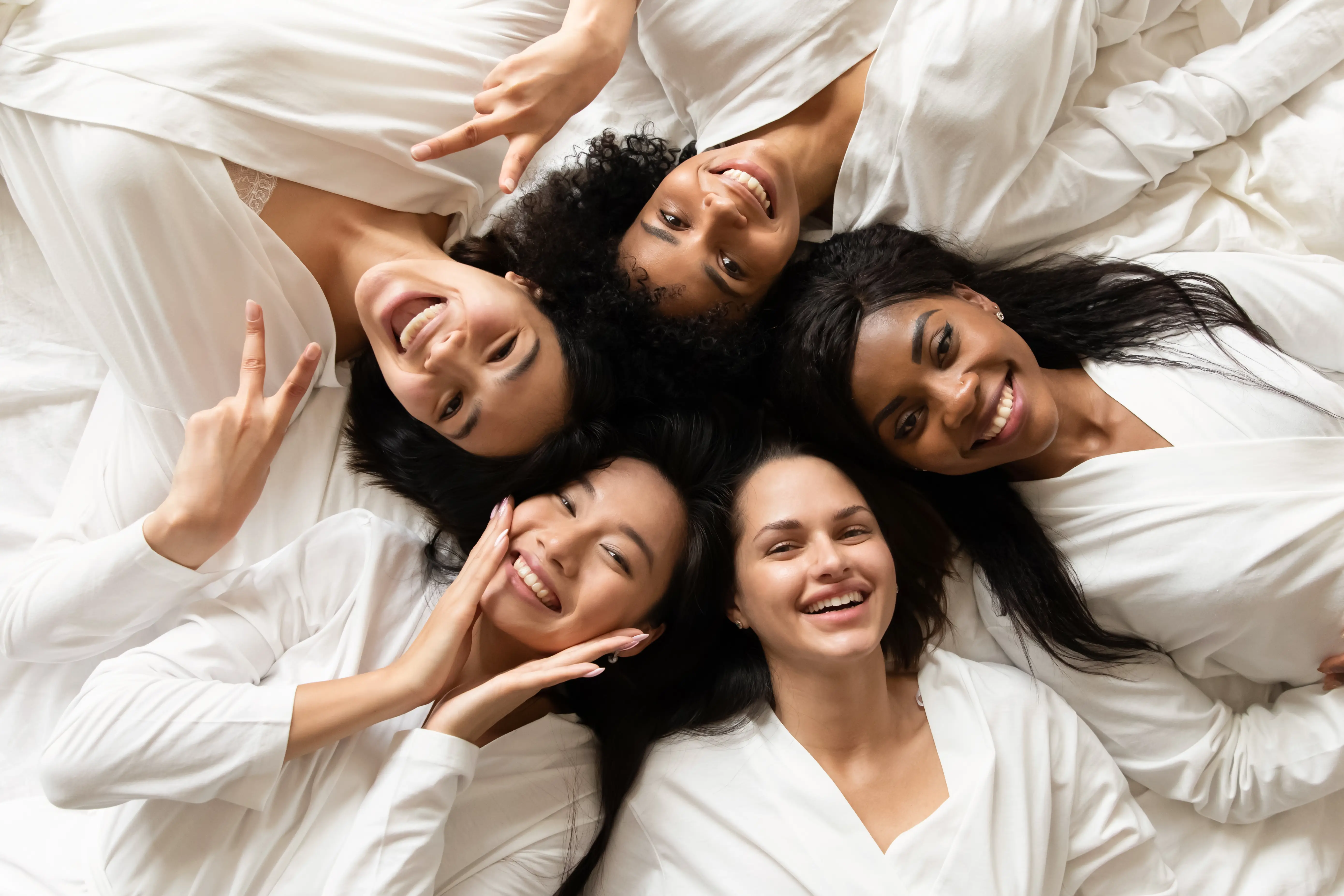 Five women lying together on white bedding and smiling at the camera
