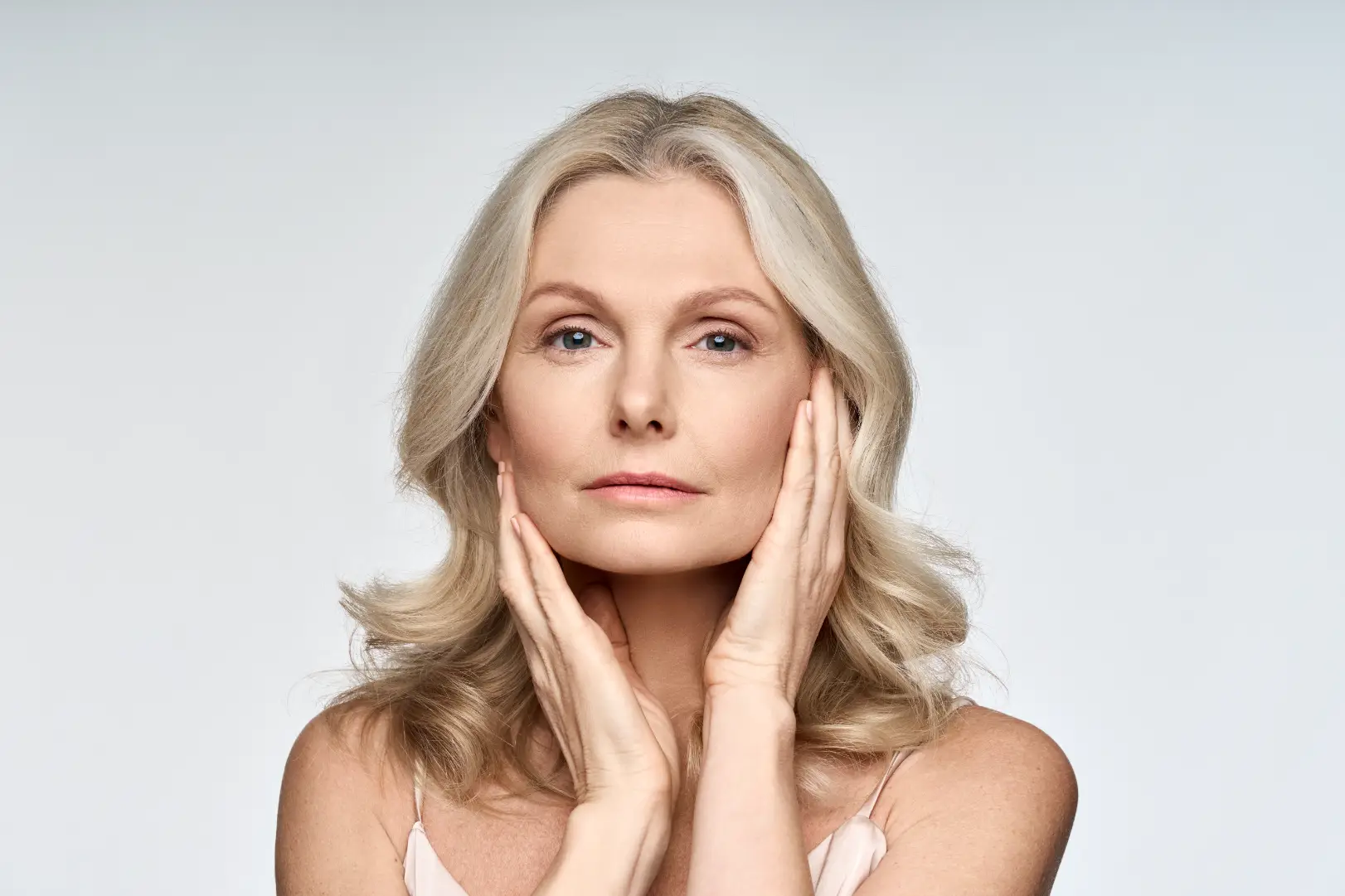 Mature blonde woman facing forward with both hands resting gently along her cheeks and jawline.