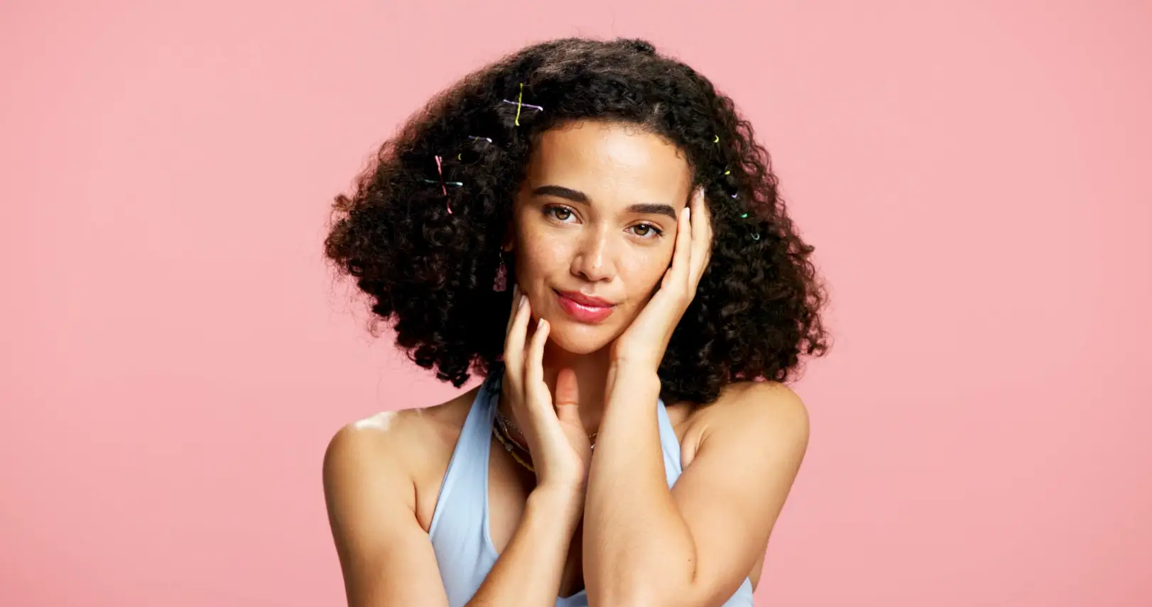 Woman with curly dark hair gently touches both sides of her face against a solid pink background.